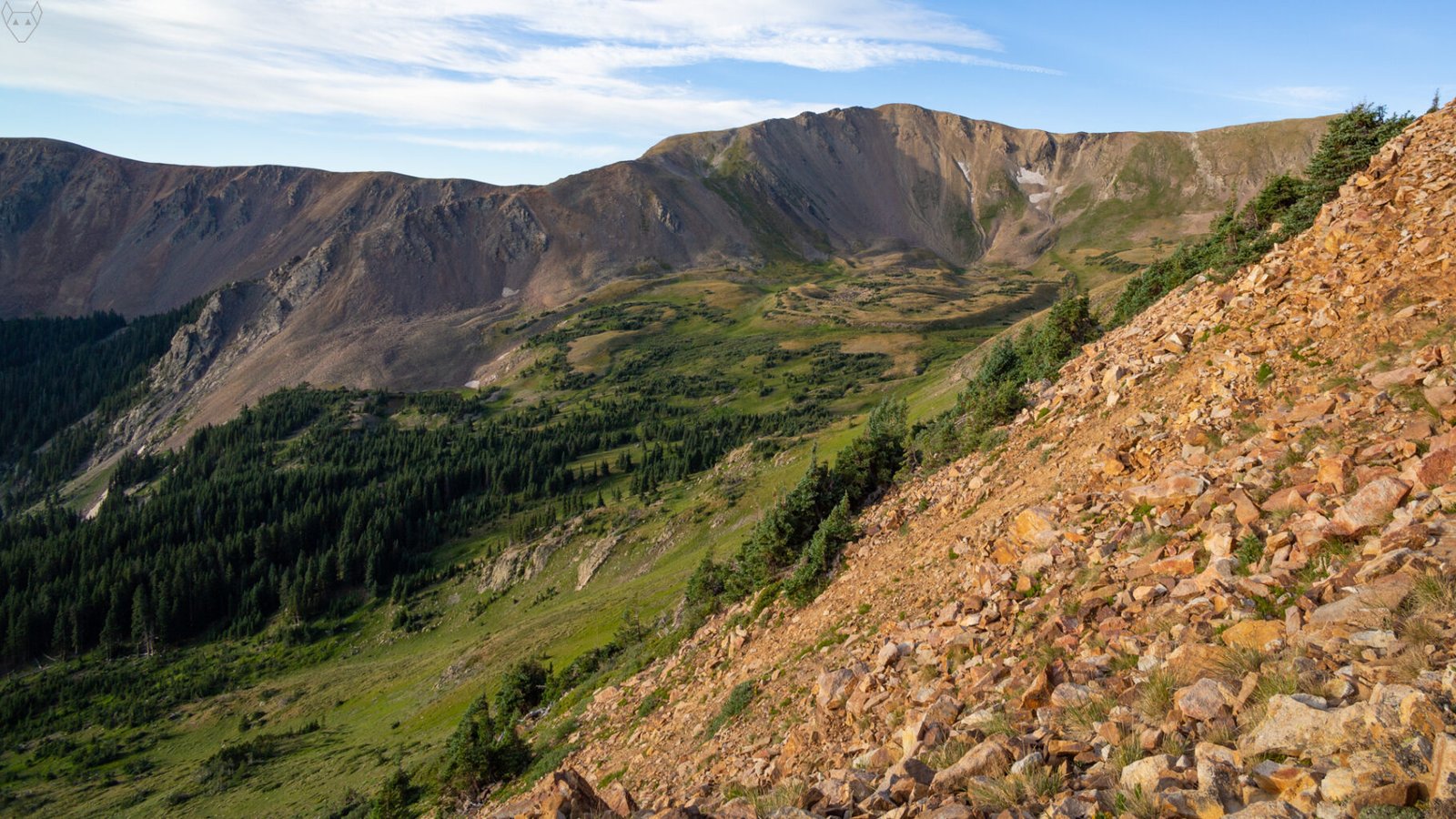 A wide mountain basin opens beneath a blue sky streaked with white clouds. A rust-colored scree slope fills the right foreground, dropping toward dark evergreen forest, green alpine meadows, rocky ridges, and small patches of lingering snow.