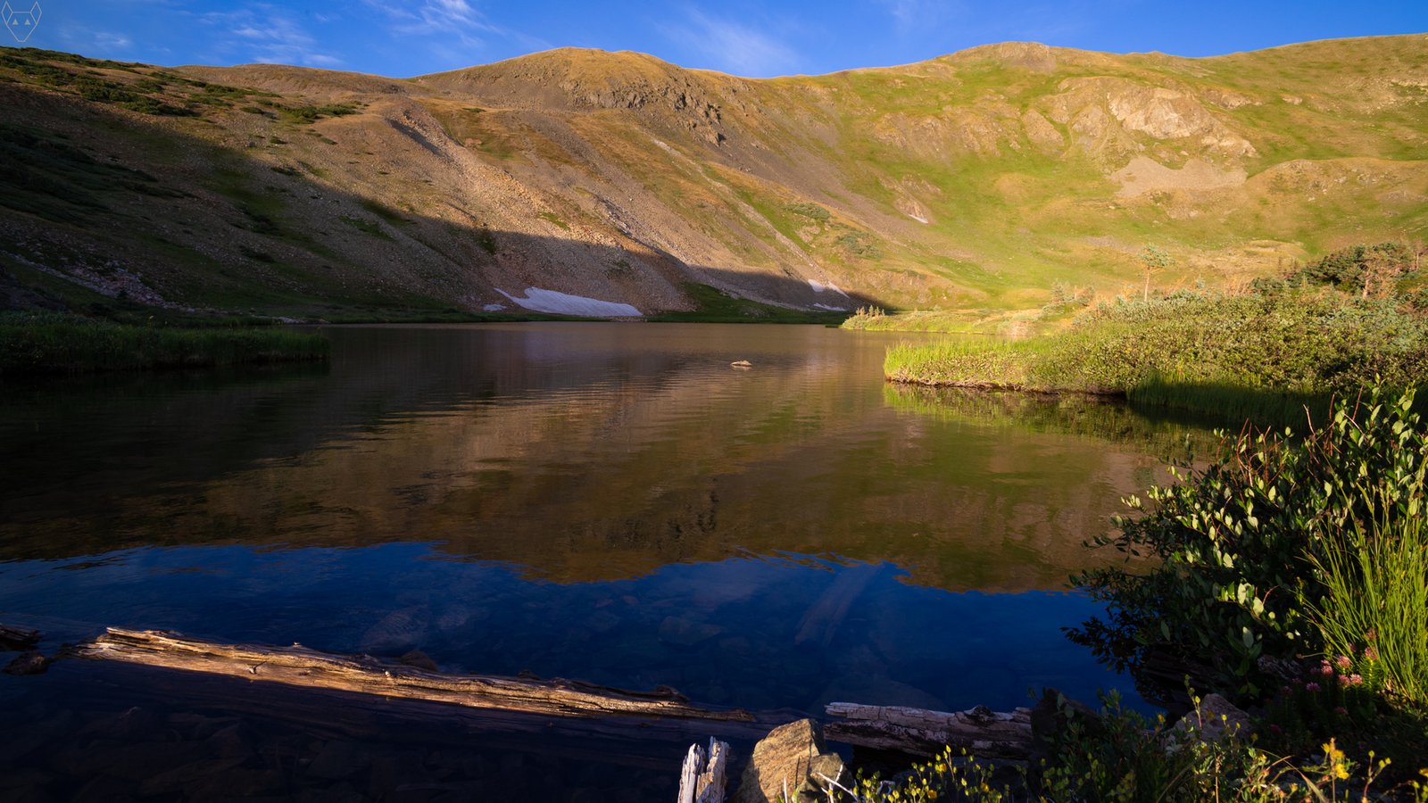 Morning light spreads across steep alpine slopes above Horseshoe Lake, New Mexico. A looming mountain shadow cuts across the water and lower basin, while golden ridges, green patches, snow remnants, and wispy clouds reflect in the still surface.