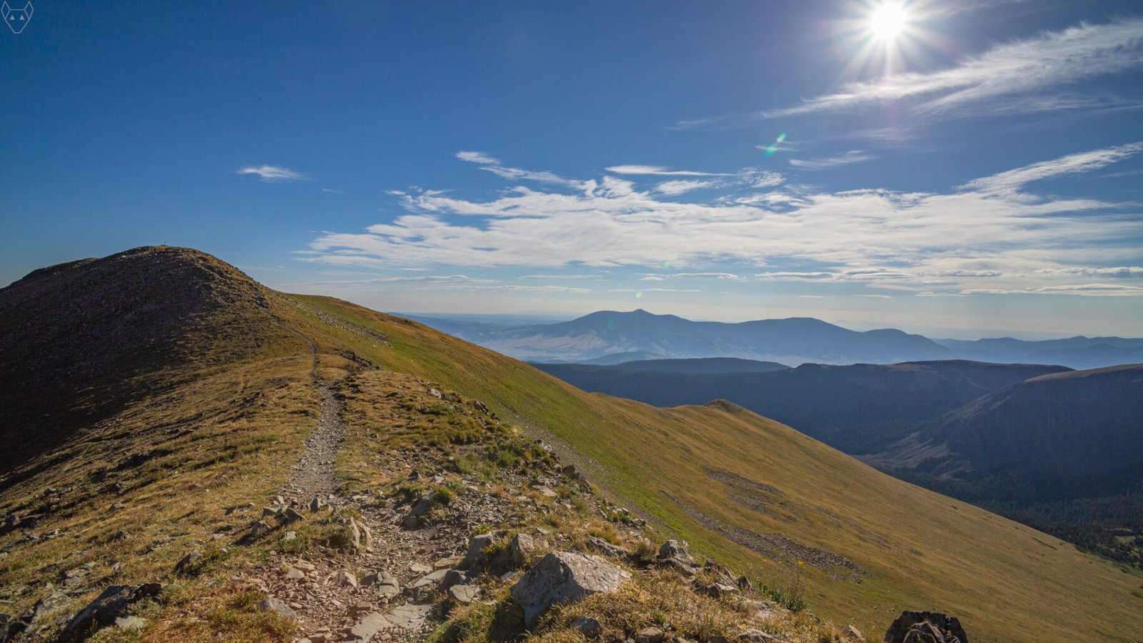 A rocky alpine trail follows a rounded ridge toward a shadowed summit under a bright sun. Golden tundra slopes fall away on both sides, with layered blue mountains and thin white clouds stretching across the horizon.