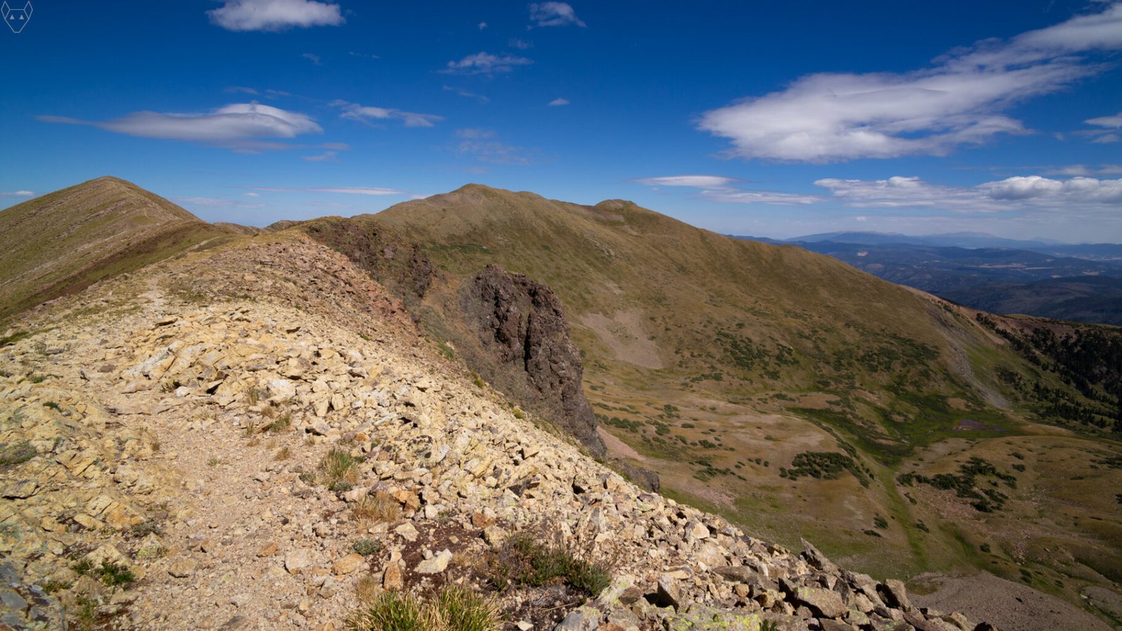A rocky alpine ridge curves across the foreground toward a chain of rounded high peaks under a deep blue sky. Steep green and brown slopes fall into a wide basin below, with dark forest, distant blue mountains, and scattered white clouds beyond.