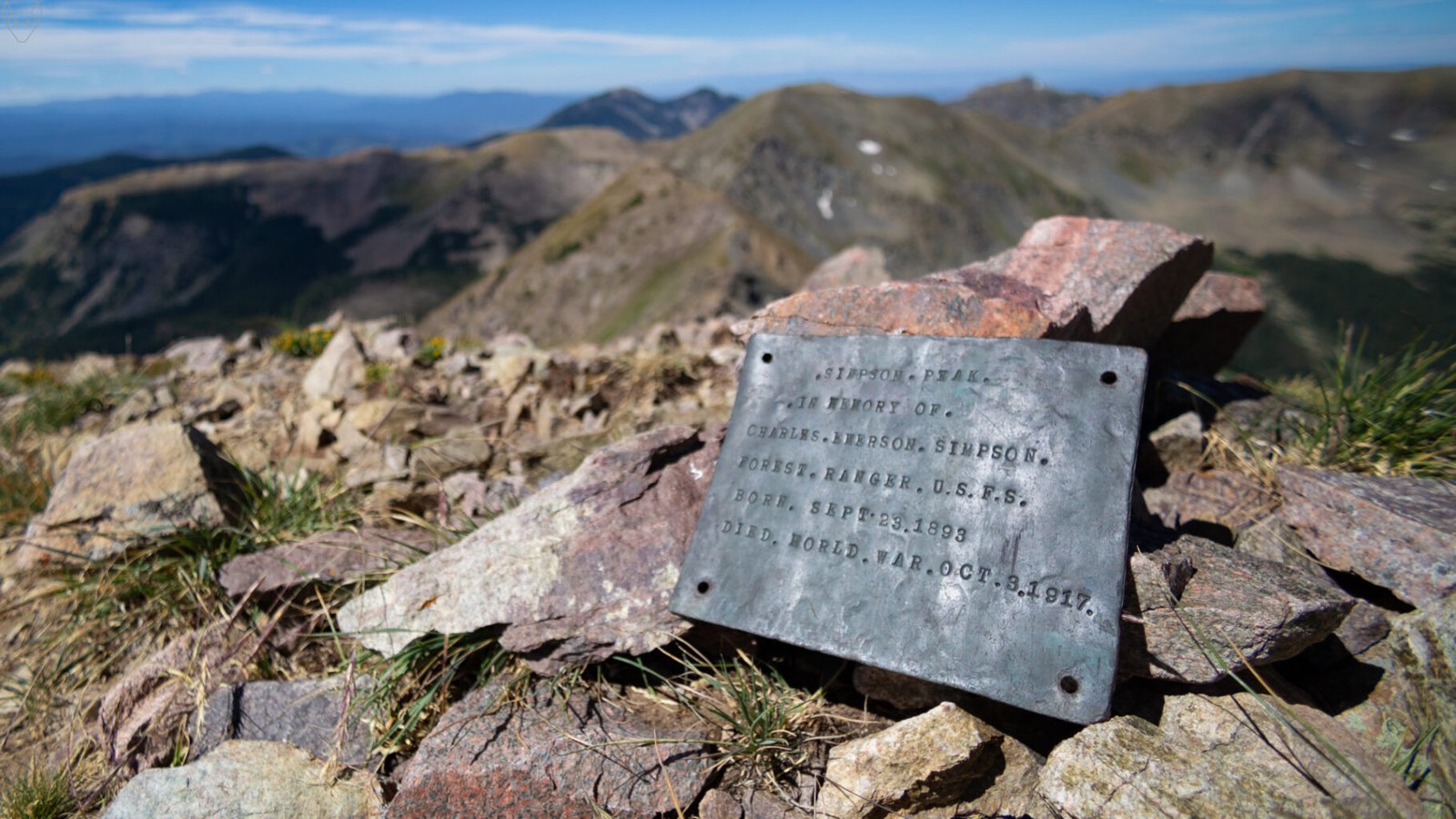 A plaque rests among pink and gray summit rocks, with blurred alpine peaks and blue distance behind it. The inscription names Simpson Peak in memory of Charles Everson Simpson, a U.S. Forest Service ranger who died in the World War on October 3, 1917.