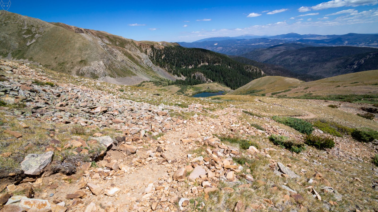 A rocky downhill trail drops through orange and gray scree toward Horseshoe Lake far below. A dark conifer forest fills the middle distance, with rounded alpine slopes, broad valleys, and layered blue mountains stretching to the horizon.
