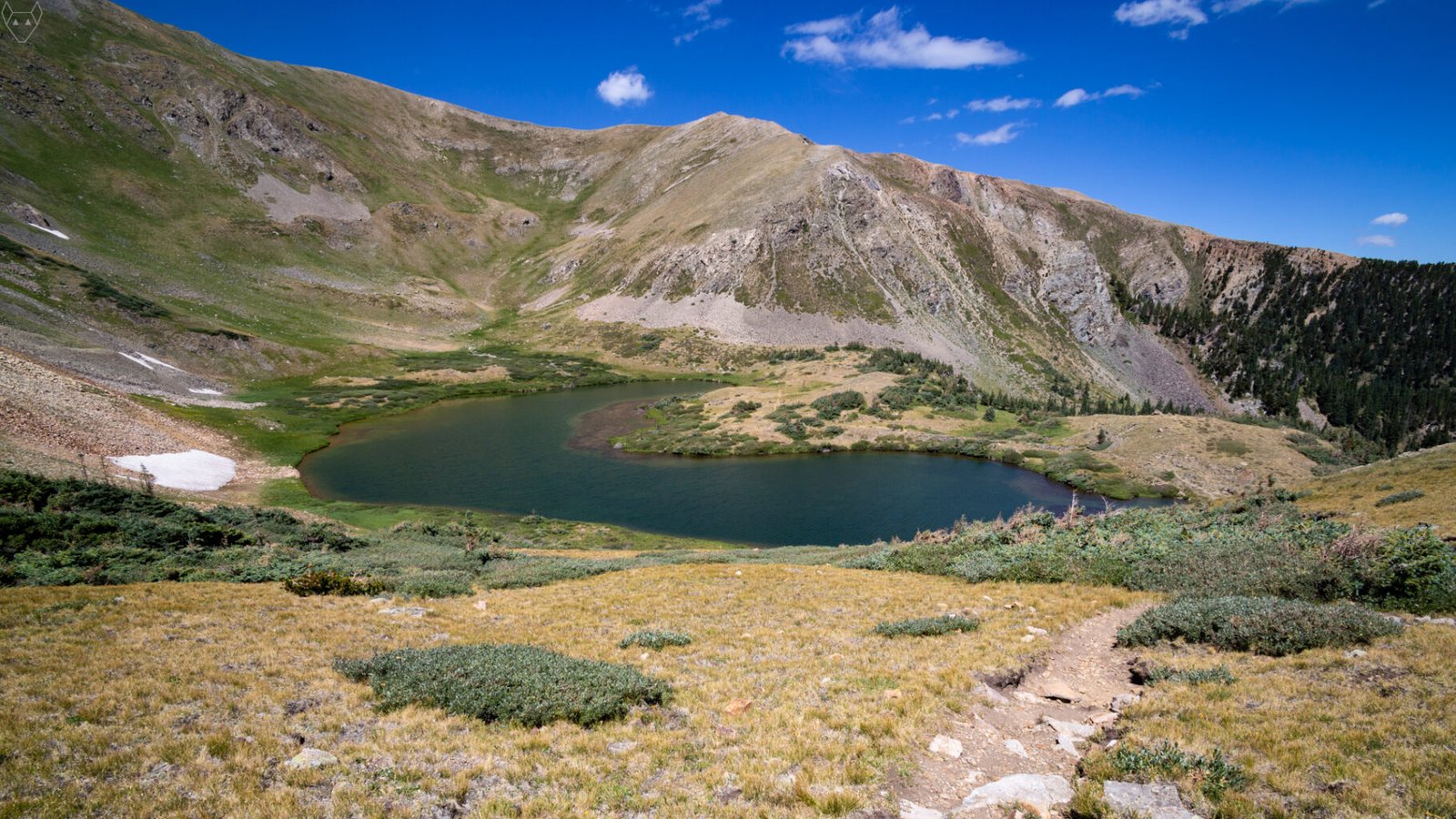 Horseshoe Lake curves through a high alpine basin below steep green and gray slopes. A small peninsula reaches into the dark blue water, with low shrubs, lingering snow, scree fields, a rocky trail, and dark conifers under a clear summer sky.