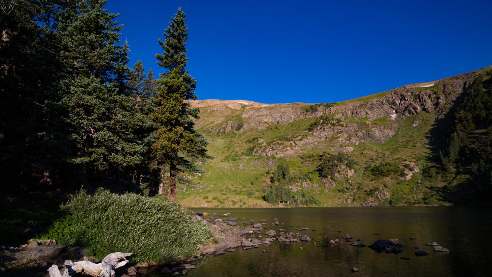 As the sun rises, Lost Lake is still and silent below steep green slopes and rocky cliffs under a cloudless deep blue sky. One Two Three A conifer forest frames the left shore, scattered stones sit in the shallow water, and a bleached fallen branch curves across the foreground.